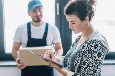 A delivery person holds a box while a woman signs a document on a clipboard.