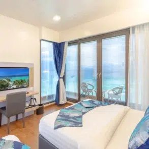 Modern hotel room with a bed, desk, TV, and a large window overlooking a turquoise ocean with balconies.