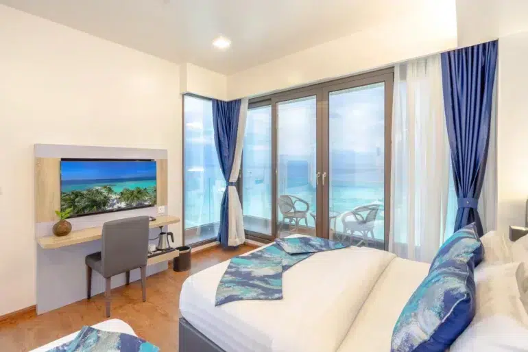 Modern hotel room with a bed, desk, TV, and a large window overlooking a turquoise ocean with balconies.