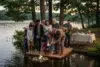 A multi-generational family stands on a wooden raft by a lake, participating in a memorial ceremony with a flower wreath and lit candles.