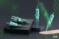 Three green and clear tattoo needle cartridges are displayed on black pedestals against a dark background.
