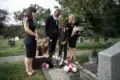 A family in black attire gathers somberly around a grave adorned with flowers in a cemetery.