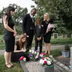 A family in black attire gathers somberly around a grave adorned with flowers in a cemetery.