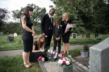 A family in black attire gathers somberly around a grave adorned with flowers in a cemetery.