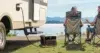 Couple sitting on camping chairs by a lake, next to an RV powered by a portable generator on the grass.
