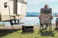 Couple sitting on camping chairs by a lake, next to an RV powered by a portable generator on the grass.