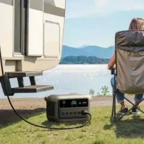 Couple sitting on camping chairs by a lake, next to an RV powered by a portable generator on the grass.
