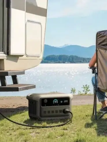 Couple sitting on camping chairs by a lake, next to an RV powered by a portable generator on the grass.