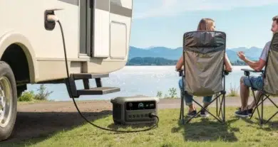 Couple sitting on camping chairs by a lake, next to an RV powered by a portable generator on the grass.