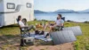 Two people relax beside a camper van at a lakeside campsite with snacks, a portable power station, and solar panels overlooking mountain scenery.