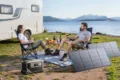 Two people relax beside a camper van at a lakeside campsite with snacks, a portable power station, and solar panels overlooking mountain scenery.