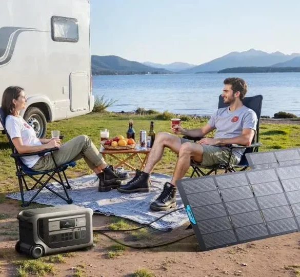 Two people relax beside a camper van at a lakeside campsite with snacks, a portable power station, and solar panels overlooking mountain scenery.