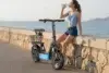 A young woman in shorts and a sleeveless top sits on a stone seawall next to a parked black electric scooter with a basket, drinking water from a bottle with the ocean in the background.
