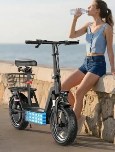 A young woman in shorts and a sleeveless top sits on a stone seawall next to a parked black electric scooter with a basket, drinking water from a bottle with the ocean in the background.