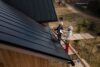 Two roofers in hard hats work on a black metal roof of a wooden building with scaffolding.