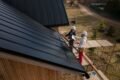 Two roofers in hard hats work on a black metal roof of a wooden building with scaffolding.