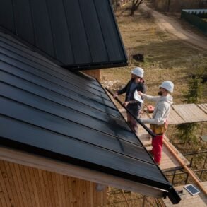 Two roofers in hard hats work on a black metal roof of a wooden building with scaffolding.