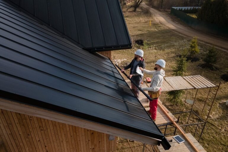 Two roofers in hard hats work on a black metal roof of a wooden building with scaffolding.