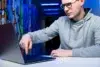 Man wearing glasses and a grey hoodie points at a laptop screen in a hallway with colorful murals and blue lighting.