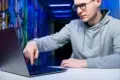 Man wearing glasses and a grey hoodie points at a laptop screen in a hallway with colorful murals and blue lighting.