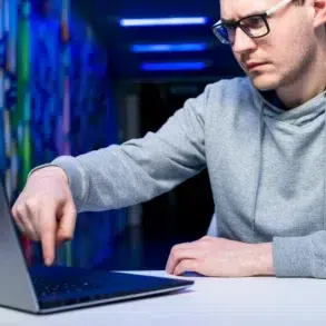 Man wearing glasses and a grey hoodie points at a laptop screen in a hallway with colorful murals and blue lighting.
