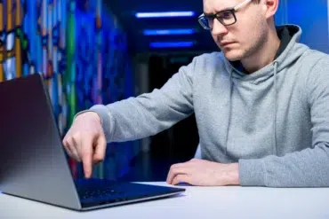 Man wearing glasses and a grey hoodie points at a laptop screen in a hallway with colorful murals and blue lighting.