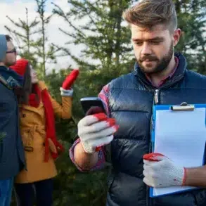 A man with a clipboard and phone stands in front of a couple choosing a Christmas tree.