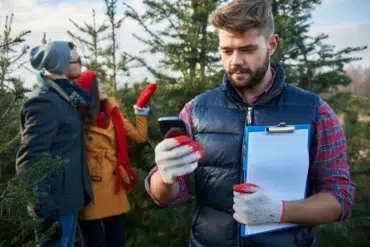 A man with a clipboard and phone stands in front of a couple choosing a Christmas tree.