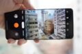 A hand holds a smartphone horizontally, displaying the camera app interface capturing an ultra-wide view of a dense urban street scene with tall buildings.