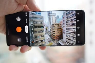 A hand holds a smartphone horizontally, displaying the camera app interface capturing an ultra-wide view of a dense urban street scene with tall buildings.