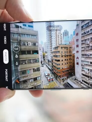 A hand holds a smartphone horizontally, displaying the camera app interface capturing an ultra-wide view of a dense urban street scene with tall buildings.