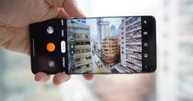 A hand holds a smartphone horizontally, displaying the camera app interface capturing an ultra-wide view of a dense urban street scene with tall buildings.