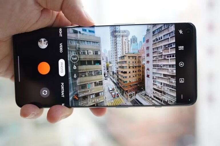 A hand holds a smartphone horizontally, displaying the camera app interface capturing an ultra-wide view of a dense urban street scene with tall buildings.