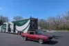 A red classic car parked in a paved lot next to a large, white and green semi-trailer with a person standing on the open ramp.