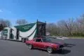 A red classic car parked in a paved lot next to a large, white and green semi-trailer with a person standing on the open ramp.