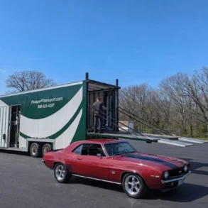 A red classic car parked in a paved lot next to a large, white and green semi-trailer with a person standing on the open ramp.
