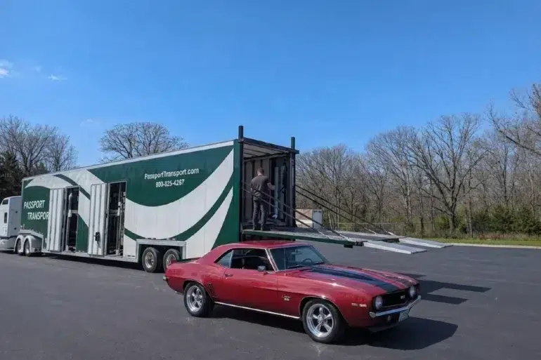 A red classic car parked in a paved lot next to a large, white and green semi-trailer with a person standing on the open ramp.