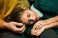 A woman with closed eyes and red lipstick rests her head on a man's shoulder, his hand gently touching her hair.