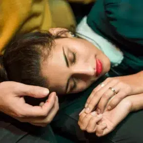 A woman with closed eyes and red lipstick rests her head on a man's shoulder, his hand gently touching her hair.