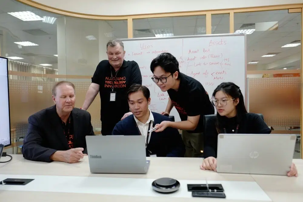 Diverse team of five collaborates around laptops in an office with a whiteboard.