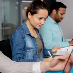 Three people working at a desk in an office, each focused on their laptops. A woman in denim looks concentrated, creating a collaborative workspace vibe.