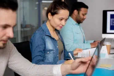 Three people working at a desk in an office, each focused on their laptops. A woman in denim looks concentrated, creating a collaborative workspace vibe.