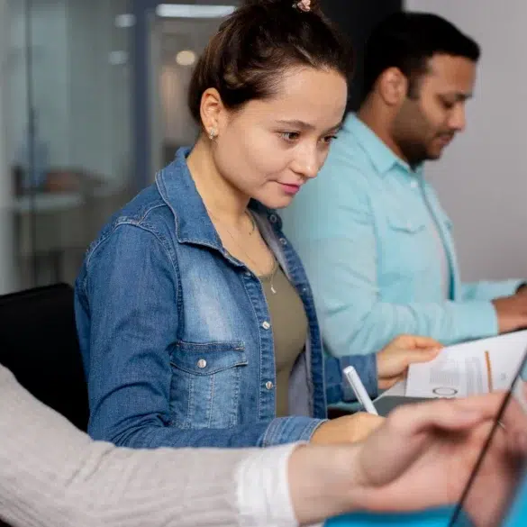 Three people working at a desk in an office, each focused on their laptops. A woman in denim looks concentrated, creating a collaborative workspace vibe.
