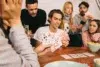 A group of friends sits around a table playing cards, displaying focus and enjoyment. Some hold hands of cards, while snacks and a drink are nearby.