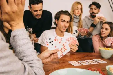 A group of friends sits around a table playing cards, displaying focus and enjoyment. Some hold hands of cards, while snacks and a drink are nearby.