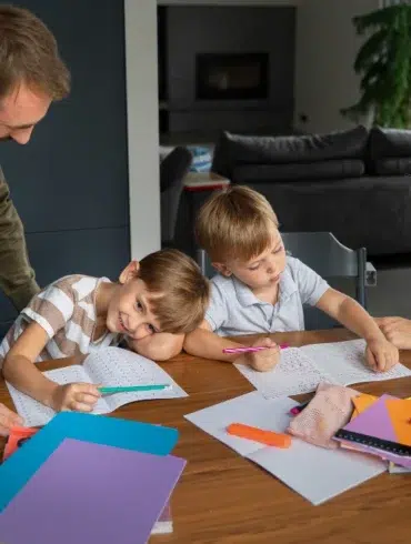 Parents help two young children with homework at a table with notebooks and school supplies.