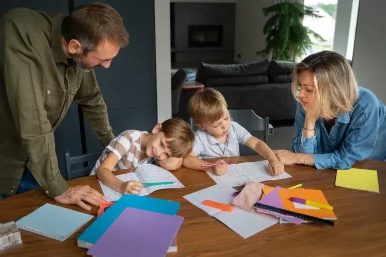 Parents help two young children with homework at a table with notebooks and school supplies.