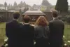Four people in dark clothing stand facing a casket at a graveside with houses in the background.