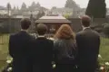 Four people in dark clothing stand facing a casket at a graveside with houses in the background.