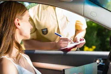 A woman in a car receiving a traffic ticket from a police officer writing on a clipboard outside the driver's window.